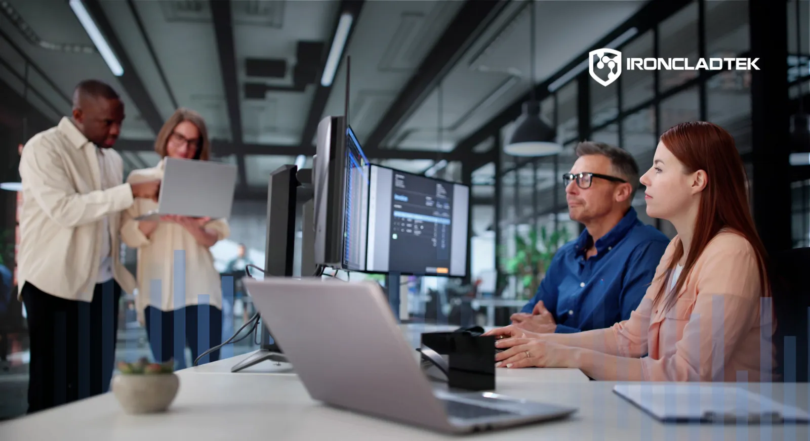 An image that represents the title of the blog: What Do Managed IT Services Cost? There is a group of people in a business. Two are standing, looking at a laptop together, while other two are seated at a large table covered with laptops and papers.